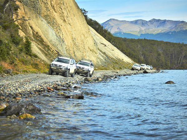 White SUVs parked on a rocky beach beside a pristine lake with snow-capped mountains and steep cliffs in Patagonia.