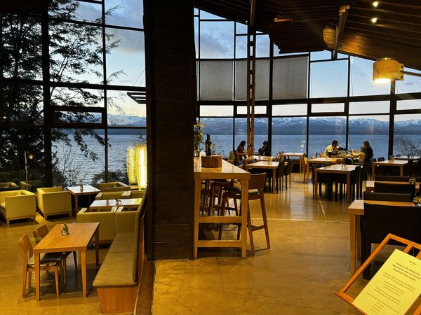 Modern lakeside restaurant with floor-to-ceiling windows overlooking Nahuel Huapi Lake near Bariloche, featuring contemporary wood and yellow furnishings.