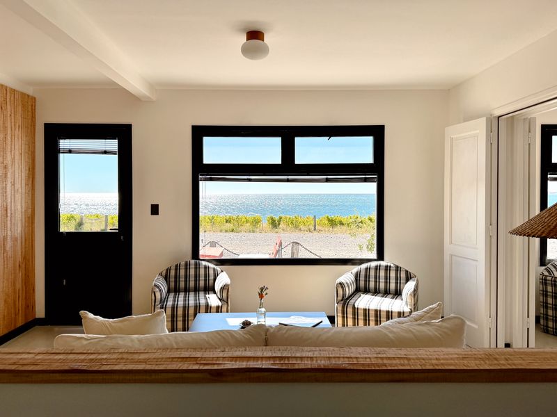 Bahía Bustamante suite with panoramic ocean views, striped armchairs, and coastal beach landscape visible through large windows.