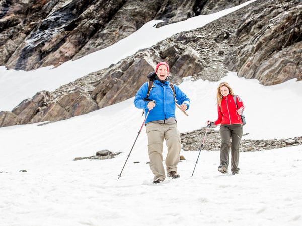 Two hikers trek across snowy Glaciar Martial in Patagonia, with dramatic rocky peaks and glacial terrain in the background.