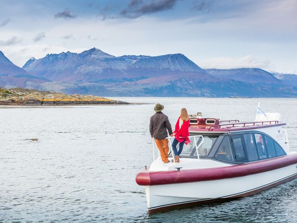 Couple standing on a boat with snow-capped mountains across the water in Lapataia, Tierra del Fuego.