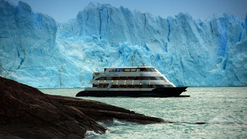 Luxury cruise ship anchored before the towering blue ice walls of Perito Moreno Glacier in Patagonia.
