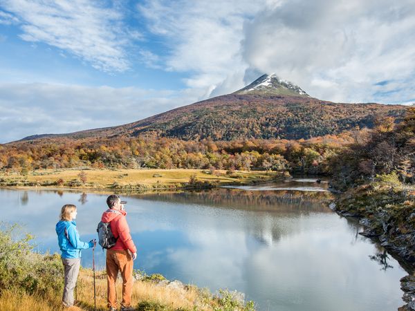 Hikers pause by a pristine lake with snow-capped mountain peaks and autumn forests in Tierra del Fuego National Park.