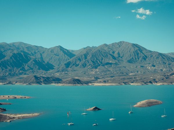Turquoise waters of Potrerillos Reservoir with sailboats, desert mountains, and arid Andean landscape under clear blue sky.