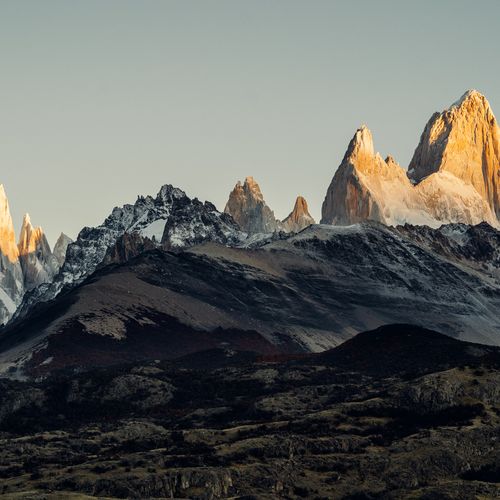 Mount Fitz Roy and Cerro Torre rise dramatically at sunrise in El Chaltén, Patagonia, with snow-capped peaks glowing golden above dark volcanic terrain.