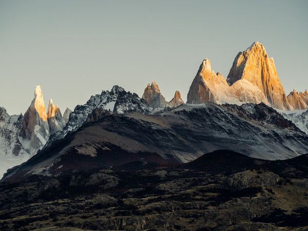 Mount Fitz Roy and Cerro Torre rise dramatically at sunrise in El Chaltén, Patagonia, with snow-capped peaks glowing golden above dark volcanic terrain.