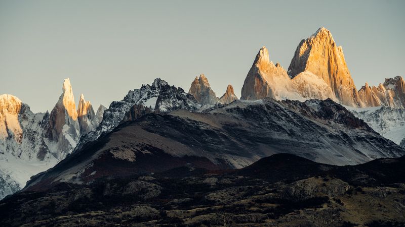 Mount Fitz Roy and Cerro Torre rise dramatically at sunrise in El Chaltén, Patagonia, with snow-capped peaks glowing golden above dark volcanic terrain.
