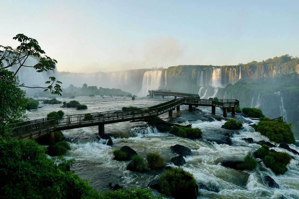 Wooden walkway extending over turbulent rapids with massive Iguazú Falls cascading in the background, surrounded by lush subtropical vegetation.