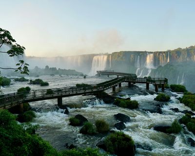 Wooden walkway extending over turbulent rapids with massive Iguazú Falls cascading in the background, surrounded by lush subtropical vegetation.
