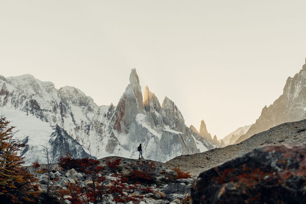 Hiker trekking toward dramatic granite peaks of Mount Fitz Roy in El Chaltén, Patagonia, with snow-capped mountains and autumn foliage.