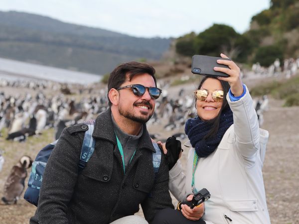 Couple taking a selfie among Magellanic penguins in Patagonia with mountains and lake in the background.