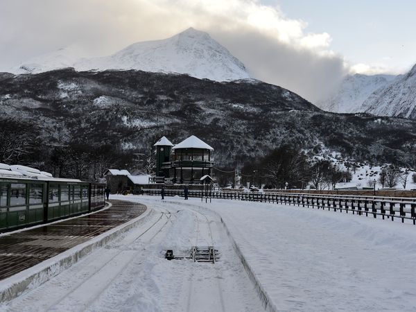 Snow-covered landscape at Tren Fin del Mundo station in Ushuaia with snow-capped mountains and rustic buildings in winter.