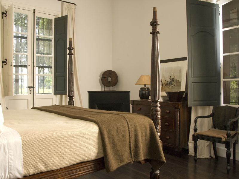 Elegant bedroom at La Bamba de Areco estancia featuring a four-poster bed, dark wood furnishings, and large windows overlooking the Buenos Aires countryside.