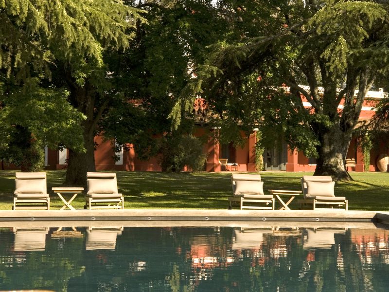 Luxurious poolside lounge at La Bamba de Areco near Buenos Aires, with cream loungers, shade trees, and a colonial red building reflected in still water.