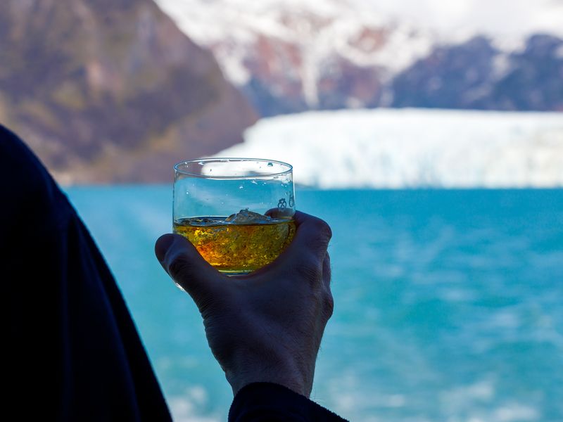 Traveler holding whisky glass with turquoise glacial lake and snow-capped mountains in El Calafate, Patagonia.