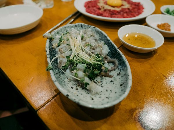 A plate of Sannakji (chopped live octopus) with greens and sesame, alongside Yukhoe and dipping sauces on a wooden table.