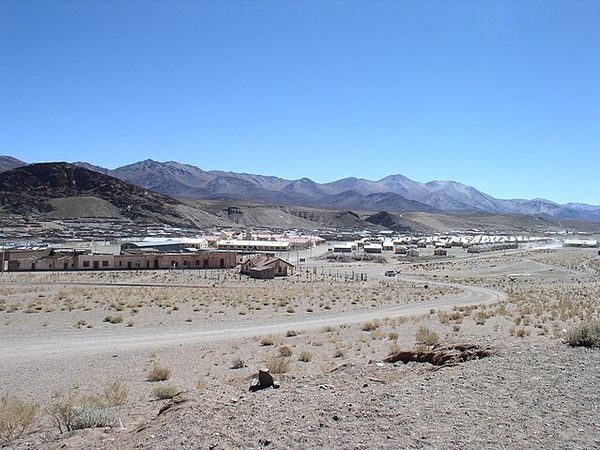Arid high-altitude town of San Antonio de los Cobres nestled in a desert valley beneath snow-capped Andean mountains under clear blue sky.