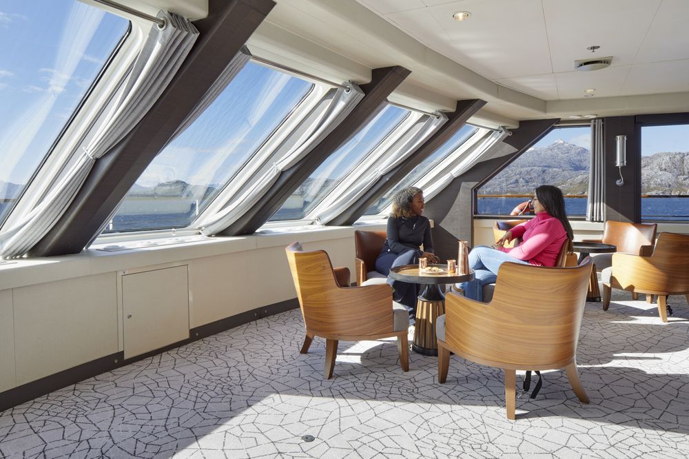 Passengers relax in a bright lounge with panoramic windows overlooking Patagonian fjords aboard an Antarctica cruise ship.