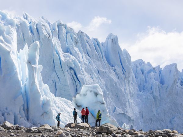 Hikers in colorful jackets stand on rocky terrain before towering ice formations at Glaciar Perito Moreno in Patagonia.