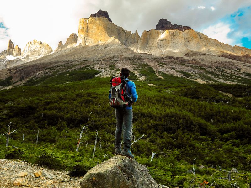 Hiker with backpack stands on rock overlooking lush valley with dramatic granite peaks of Torres del Paine, Patagonia.