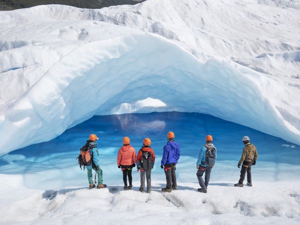 Group of hikers in colorful jackets stand before a massive blue ice cave carved beneath towering white glacial formations in Patagonia.