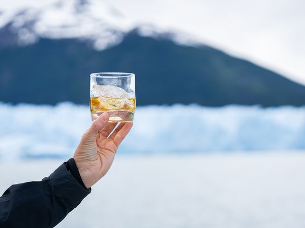 Hand holding whiskey glass with ice against snowy Patagonian glacier and mountain backdrop.