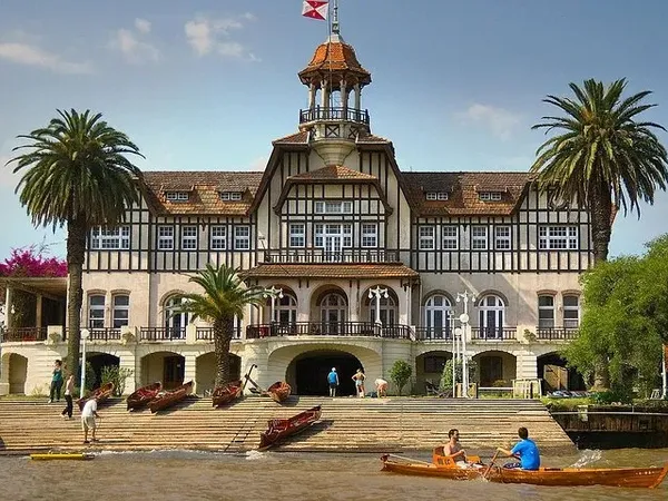 Victorian mansion with tower overlooks the Tigre River delta in Buenos Aires, surrounded by palm trees and wooden boats.