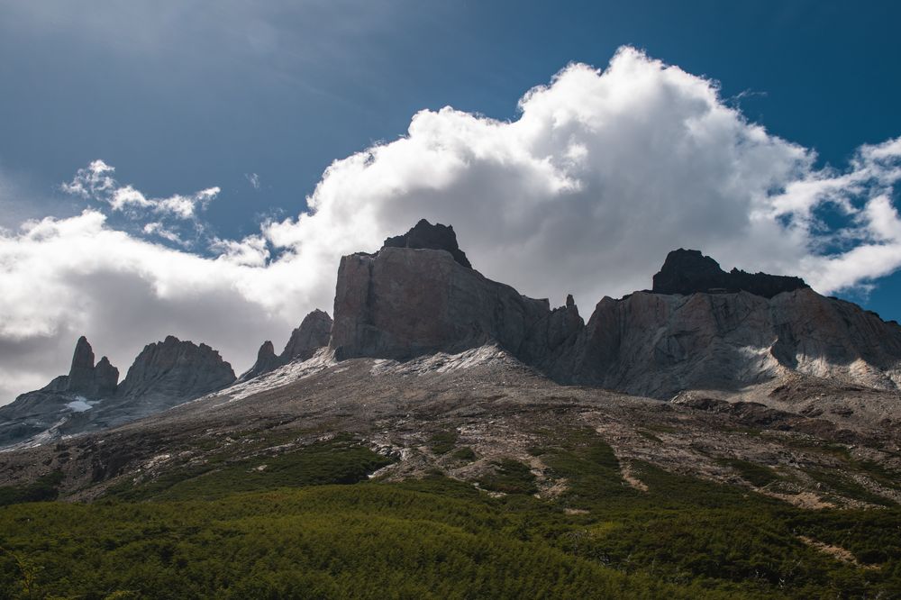 Dramatic granite peaks of Los Cuernos rise above forested slopes under a bright blue sky with white clouds in Torres del Paine, Patagonia.