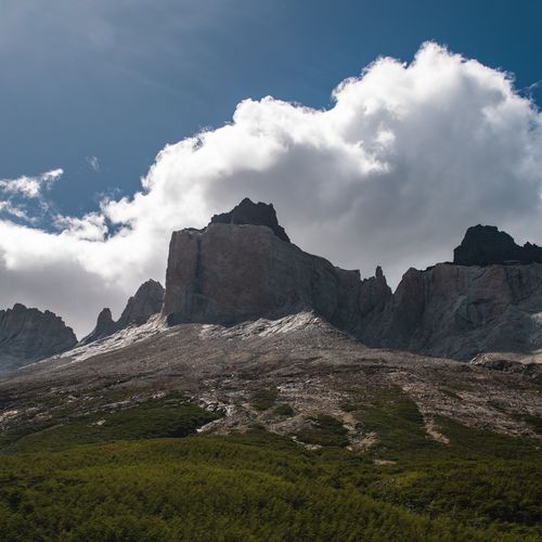 Dramatic granite peaks of Los Cuernos rise above forested slopes under a bright blue sky with white clouds in Torres del Paine, Patagonia.