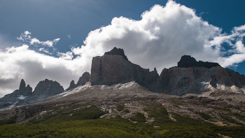 Dramatic granite peaks of Los Cuernos rise above forested slopes under a bright blue sky with white clouds in Torres del Paine, Patagonia.