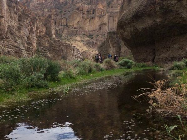 Hikers exploring a river canyon with towering rock cliffs and ancient cave dwellings carved into the mountainside in Patagonia.