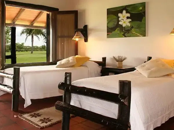 Bright twin bedroom at Aguapé Lodge with black wooden beds, green field views through open doors, and warm lighting.