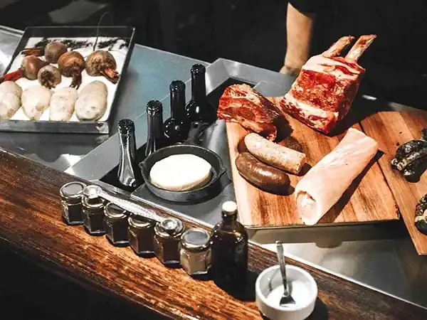 Butcher's counter with raw beef cuts, sausages, potatoes, beets, charred peppers, and spices ready for cooking.