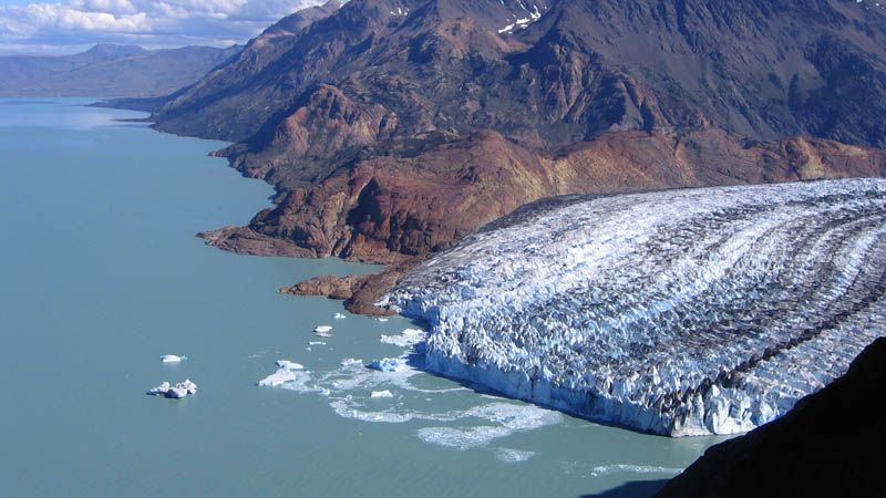 Panoramic view of the huge blue-white Perito Moreno glacier meeting a turquoise lake with icebergs, framed by rocky mountains.