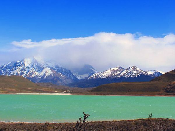 Turquoise Laguna Amarga with snow-capped peaks of Torres del Paine under cloudy Patagonian sky.
