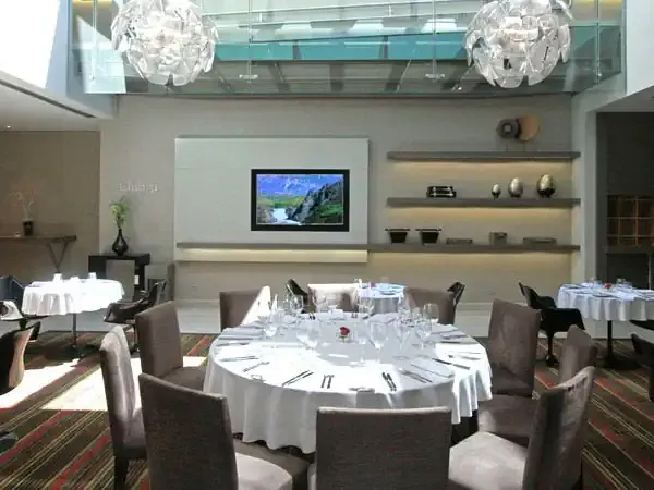 Elegant dining room at Fierro Hotel Buenos Aires with round tables, modern chandeliers, and mountain landscape on display screen.