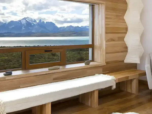 Minimalist wooden lodge interior with panoramic window overlooking turquoise lake and snow-capped Patagonian mountains.
