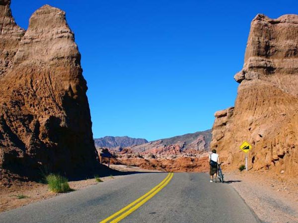 Cyclist riding through dramatic red rock formations along Ruta 9 in Cafayate's Quebrada gorge under clear blue sky.