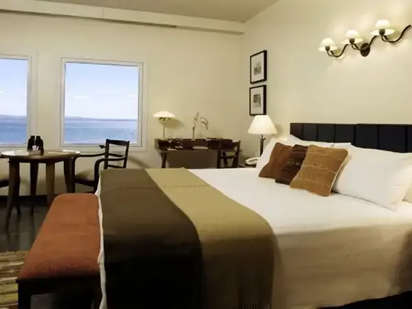 Luxury hotel bedroom with ocean views, featuring a king bed, sitting area, and panoramic windows overlooking Patagonian waters.