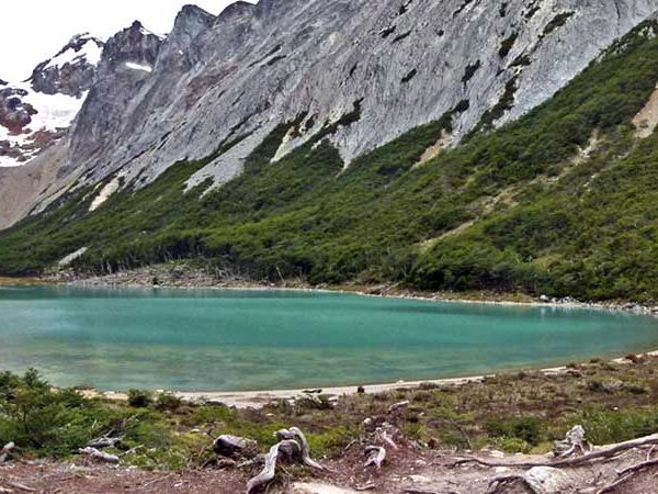 Vibrant turquoise glacial Esmeralda lagoon surrounded by green mountain slopes and distant snow-capped peaks under an overcast sky.