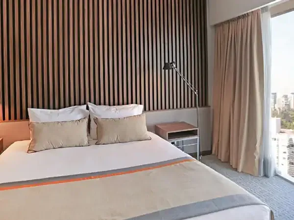 Modern hotel bedroom with city skyline view, featuring a queen bed with orange accent stripe and floor-to-ceiling windows.