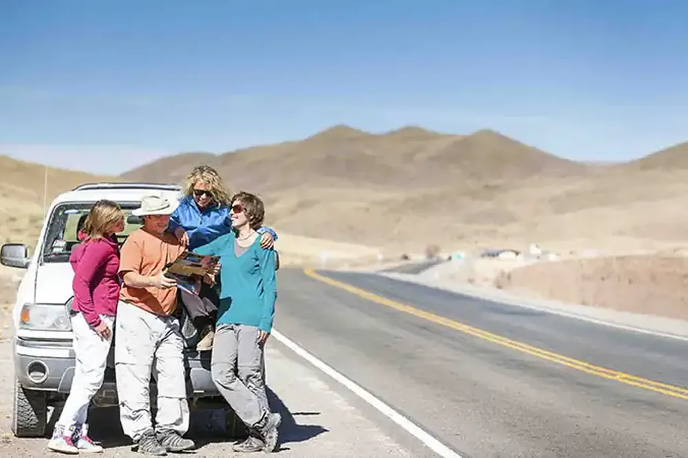 Four friends with a truck, looking at a map on a desert road with mountains.