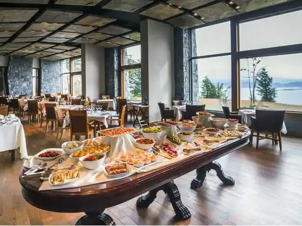 Elegant dining room at Arakur Ushuaia with gourmet buffet, floor-to-ceiling windows overlooking the Beagle Channel and mountains.