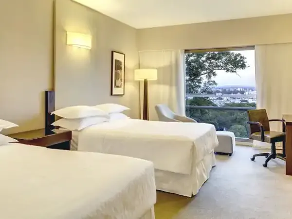 Luxury hotel room at Sheraton with twin beds, modern furnishings, and panoramic city views through floor-to-ceiling windows.