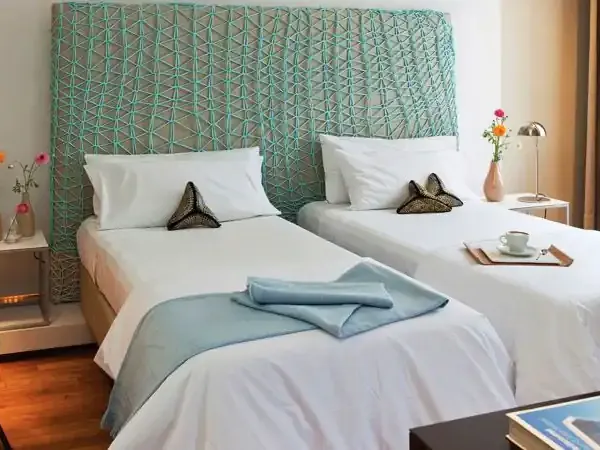 Modern hotel twin bedroom with turquoise geometric headboard, white linens, fresh flowers, and warm wood flooring.