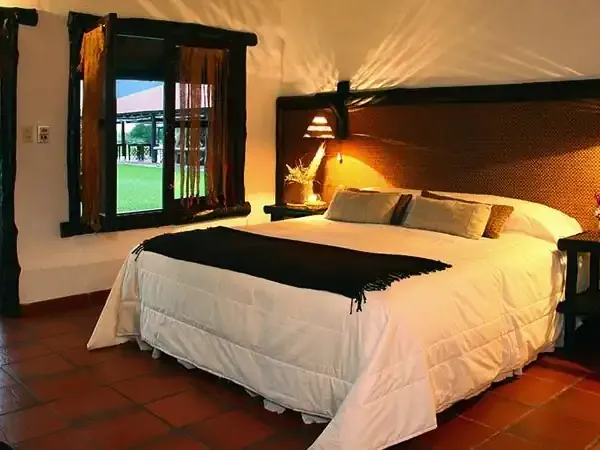Luxurious bedroom at Aguapé Lodge with king bed, warm lighting, terracotta floors, and doors opening to lush Patagonian gardens.