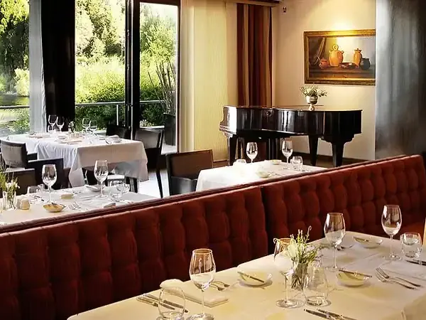 Elegant fine dining room at Casco in Patagonia with white tablecloths, red velvet banquette seating, grand piano, and lake views.