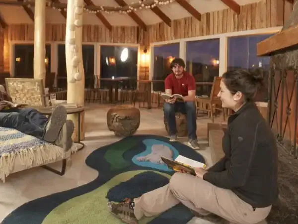 Guests relax in a rustic lodge common area at Hotel Las Torres with large windows overlooking Patagonian landscapes at dusk.