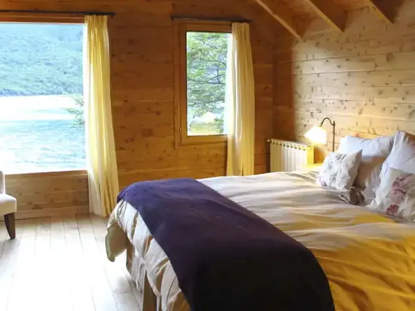Rustic wooden lodge bedroom with panoramic lake views, yellow curtains, and comfortable bedding overlooking Patagonian waters.