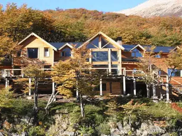 Modern wooden lodge nestled in Patagonian forest with autumn foliage and snow-capped mountains in background.